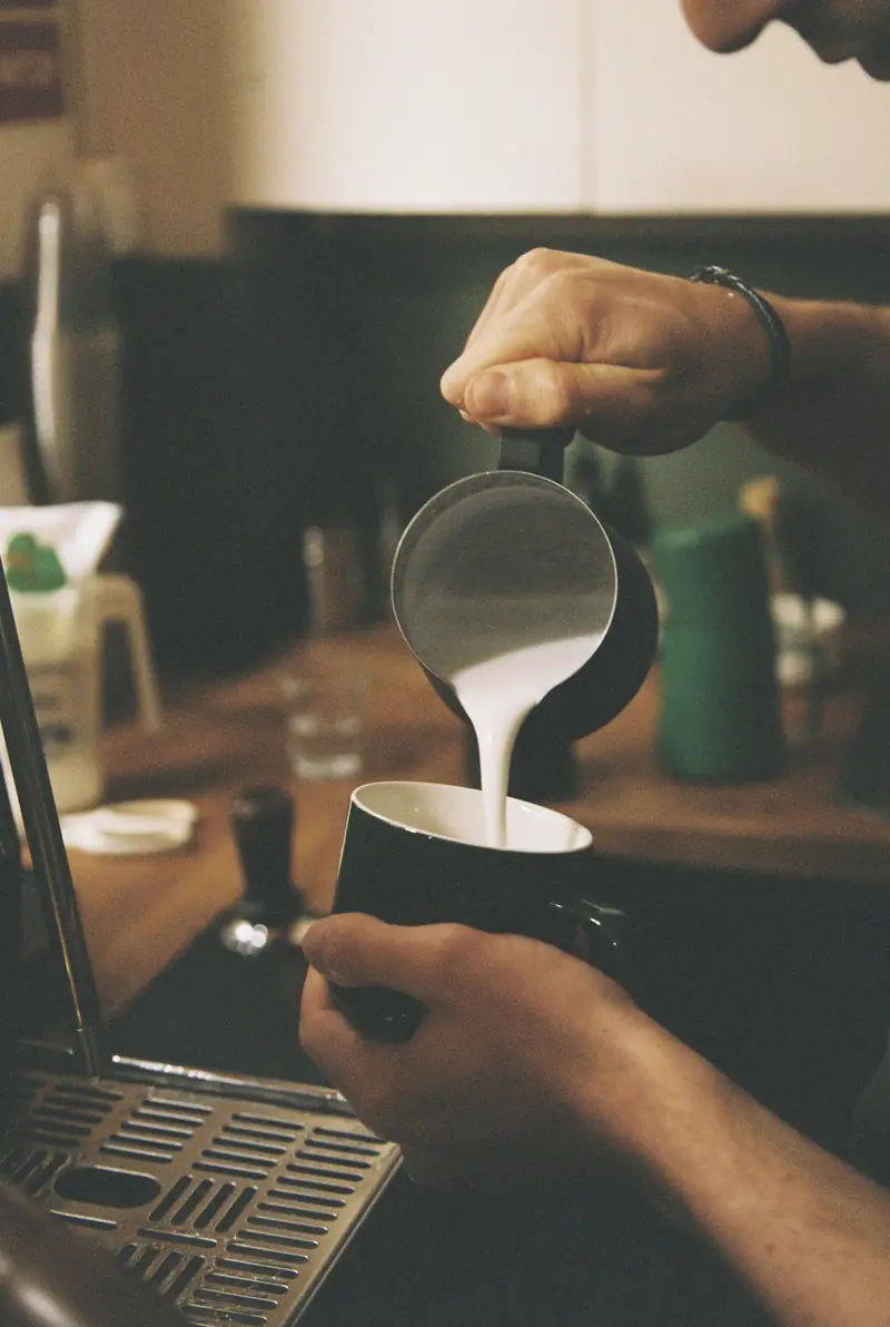 Manos de barista vertiendo leche en una jarra bajo la máquina de espresso