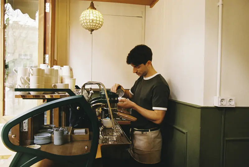 Barista trabajando en la máquina de espresso verde, con luz natural entrando por la ventana
